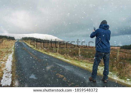 Man in winter clothes taking pictures on his smart phone on a narrow small road to a mountain during snow fall. Winter season concept. Sligo, Ireland.