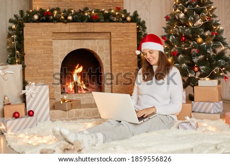 Photo of cute nice charming attractive girl having video call in eve of new year with her laptop, wearing casual white sweater and red hat, looks at device's camera with charming smile.