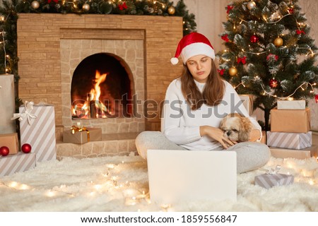 Woman and puppy dog in sweater having video call chat on laptop, enjoy Christmas time at home near x-mas tree and fireplace, hugging her favorite pekingese dog, looks sad.