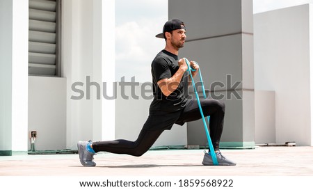 Handsome Latino sports man doing lunge workout with resitance band outdoors in the sun Royalty-Free Stock Photo #1859568922