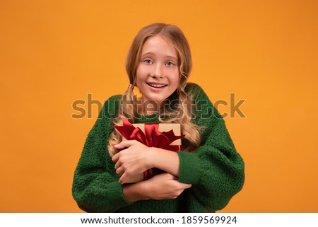 Image of charming blonde girl 12-14 years old in warm green sweater smiling and holding present box with red bow. Studio shot, yellow background. New Year Women's Day Birthday Holiday concept