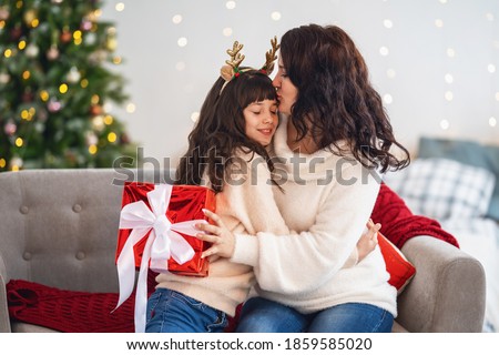 Merry Christmas and Happy Holidays! Happy mom and daughter hugging, and hold with christmas gift box on background Christmas tree decorated with a garland. Copy space