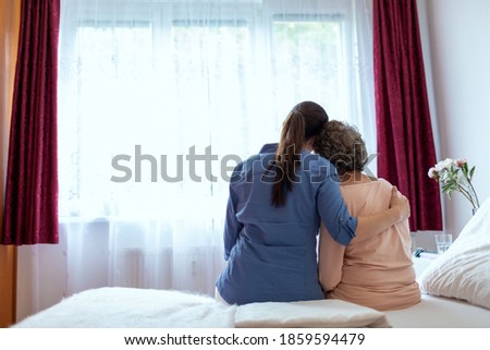 Female Home Nurse Hugging Elderly Woman on Bed. Back View of Female Nurse With Her Arm Around Elderly Patient Shoulder. Royalty-Free Stock Photo #1859594479