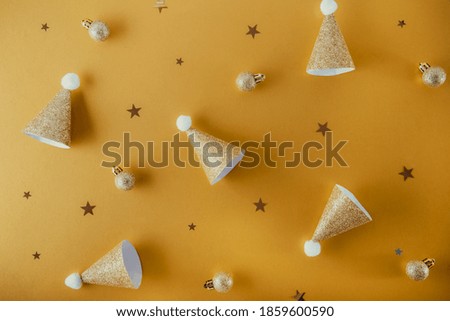 Creative Christmas background in trendy golden color. Festive paper shining caps, small balls and stars on a warm yellow background. New Year layout concept. Festive mood banner. Selective focus