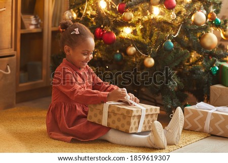 Side view portrait of happy African-American girl opening Christmas presents while sitting by tree at home, copy space
