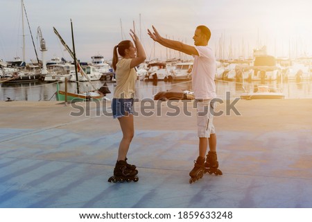 Young beautiful couple roller skating giving high five two hands at sunset in skate park next to the port in summer.Friendship lifestyle concept, success and happiness in sports competition.