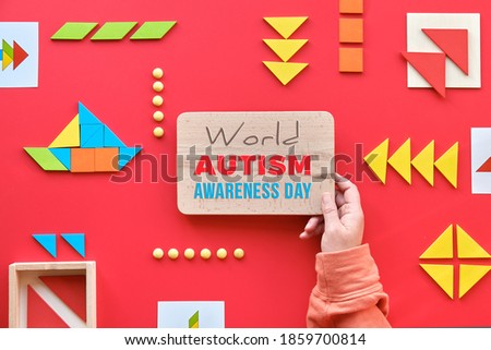 Creative design for Autism World Day on April 2. Hand hold wooden board with text World Autism Day. Tangram elements scattered and arranged in pictogram on red background.