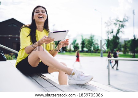 Happy ethnic female in casual clothes sitting on wooden podium in park and smiling while browsing tablet and looking at camera