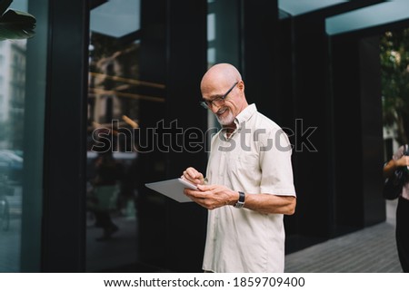 Cheerful male on retirement spending walk time for networking in city using modern gadget for browsing wireless, happy Caucaisan man in optical eyewear using touch pad for reading online news