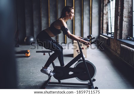 Side view of young sporty female in activewear spinning on stationary bike in loft styled gym and listening to music through earphones Royalty-Free Stock Photo #1859711575