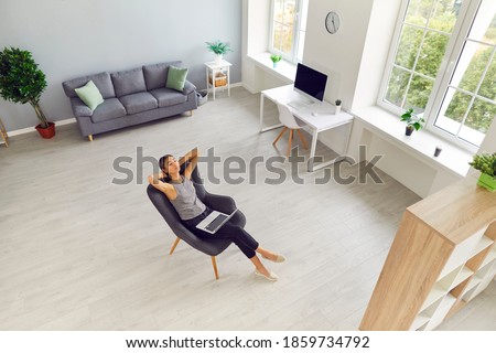 Spacious living room, study or office workspace with woman taking break from work on laptop. Modern interior of new apartment with large windows and person relaxing in comfortable armchair. High angle Royalty-Free Stock Photo #1859734792