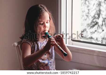 Blonde caucasian girl playing the flute netx the window at home. Empty copy space for Editor's text.
