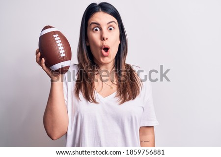 Young beautiful brunette woman playing rubgy holding football ball over white background scared and amazed with open mouth for surprise, disbelief face