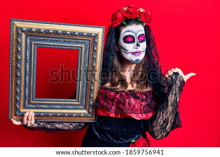 Young woman wearing day of the dead costume holding empty frame pointing thumb up to the side smiling happy with open mouth 