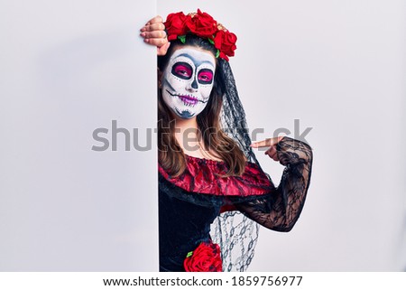 Young woman wearing day of the dead custome holding blank empty banner looking confident with smile on face, pointing oneself with fingers proud and happy. 