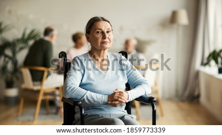 Portrait of unhappy disabled senior woman in wheelchair looking at camera with sadness in nursing home, other aged patients in background Royalty-Free Stock Photo #1859773339