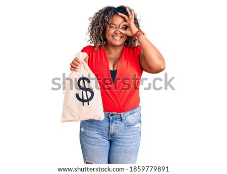 Young african american plus size woman holding money bag with dollar symbol smiling happy doing ok sign with hand on eye looking through fingers 