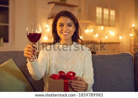 Happy young brunette woman holding glass of wine and holiday present during online Valentines day celebrating from home. Valentines day, holiday gift, anniversary, dating, distant celebration concept