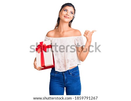 Young beautiful woman holding gift pointing thumb up to the side smiling happy with open mouth 
