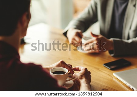 Two unrecognizable businessmen having business talk over coffee sitting at table in cafe. Laptop, coffee cups and smartphone on table. Focus on hands and cup of espresso Royalty-Free Stock Photo #1859812504