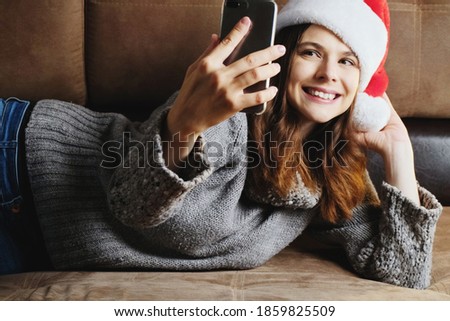Christmas selfie. Young blogger using phone, Christmas concept. Girl wearing Santa hat photographing herself smiling lying on sofa.