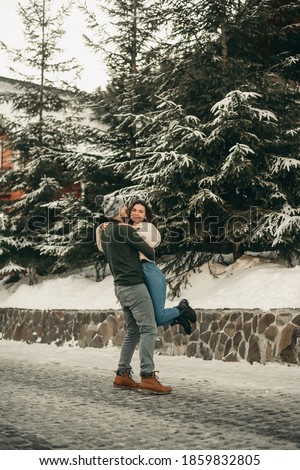 Young happy couple on the street in winter in a hat and sweater walking and having fun near a wooden house on vacation. Woman and man hugging, kissing, celebrating Christmas, ski resort, snow.