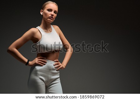 Sports blonde girl in white training clothes with headphones in the studio on a grey background. The concept of training, healthy lifestyle, portrait of a fitness instructor. Copy space.