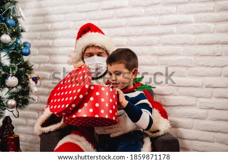 A smiling boy in a striped T-shirt who sits in Santa's lap with a face mask and opens a red xmas gift next to the Christmas tree. New Year and Christmas during the COVID-19 coronavirus pandemic