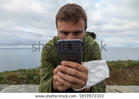 A young man, grabbing a protective face mask, holds a smart mobile phone, with the sea behind him, Spain.