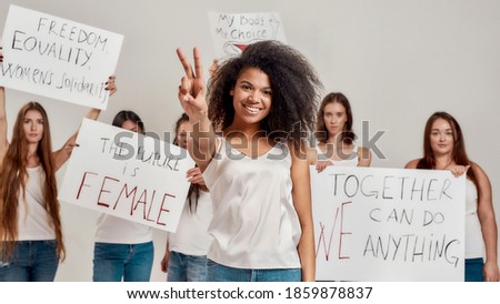 Young african american woman in white shirt showing peace sign, smiling at camera. Group of diverse women holding protest banners for woman power and rights in the background. Feminism and equality