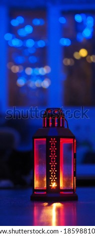 a red metal candlestick with cut patterns with glass and a burning candle inside stands on the floor against a blue background. vertical image, soft focus, space for text
