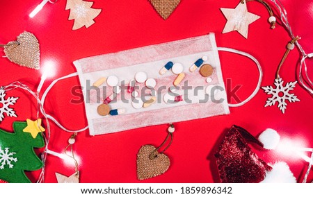 The inscription 2021 of tablets on a white protective medical mask on a red background and Christmas decorations around. Cute Santa hat, small Christmas tree and garland with bright lights.