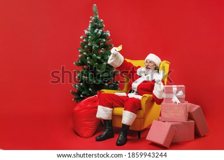 Funny Santa Claus man in Christmas suit sit in armchair with fir tree gifts doing selfie shot on mobile phone greeting with hand isolated on red background. Happy New Year celebration holiday concept