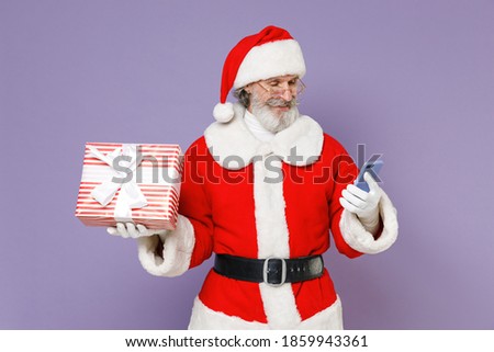 Cheerful Santa Claus man in Christmas hat red suit coat gloves glasses hold present box with gift ribbon bow isolated on violet background studio. Happy New Year celebration merry holiday concept