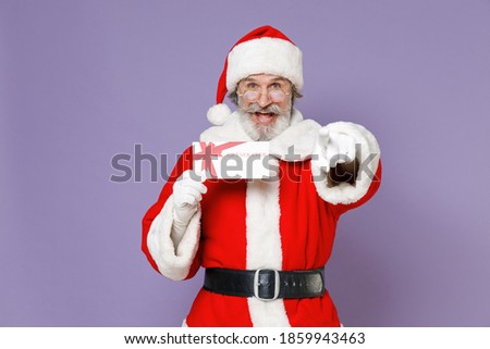 Funny Santa Claus man in Christmas hat red suit coat gloves glasses hold gift certificate point index finger on camera isolated on violet background. Happy New Year celebration merry holiday concept