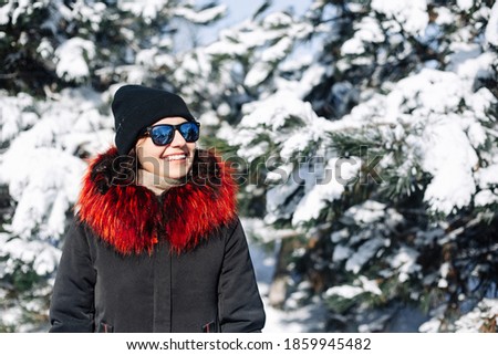 Portrait of a girl teenager wearing black and red coat standing among fir trees covered with snow at the winter park. Young woman having fun in the winter forest. Winter vibes concept.
