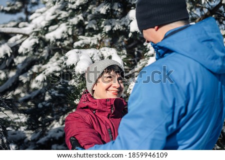 Middle age couple hug at the snowy winter park. A man and a woman in blue and red jackets having fun among fir trees covered with snow. Love, togetherness and holidays concept.