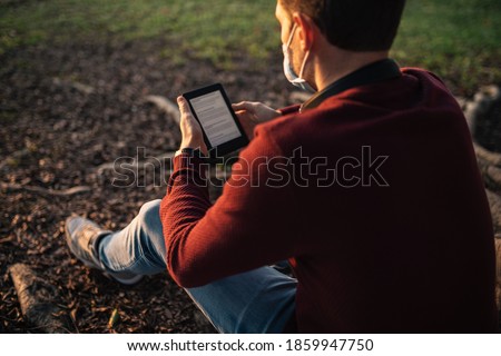 boy with a mask by the covid-19, reading an e-book, sitting in a tree at sunset. Royalty-Free Stock Photo #1859947750