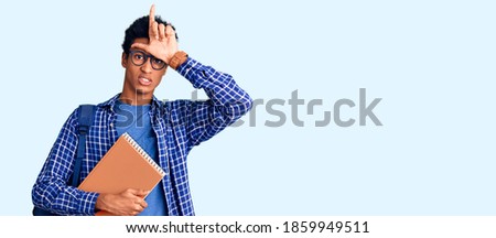 Young african american man wearing student backpack holding book making fun of people with fingers on forehead doing loser gesture mocking and insulting. 