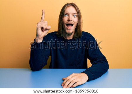 Handsome caucasian man with long hair wearing casual clothes sitting on the table pointing finger up with successful idea. exited and happy. number one. 