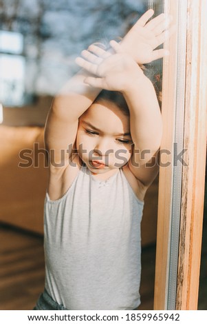 sad child stands behind the glass in the window of the house with his arms crossed during quarantine