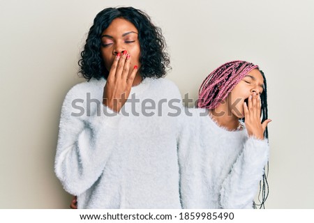 Beautiful african american mother and daughter wearing casual winter sweater bored yawning tired covering mouth with hand. restless and sleepiness. 