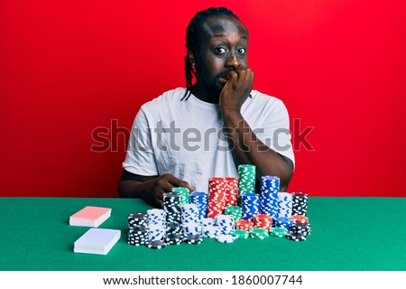 Handsome young black man sitting on the table with poker chips and cards looking stressed and nervous with hands on mouth biting nails. anxiety problem. 