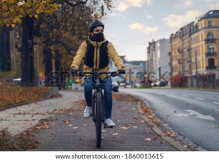 A man, wearing a winter jacket, and the mask for the virus crown / covid 19, cycling in the city. Royalty-Free Stock Photo #1860013615