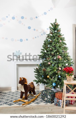 Poinsettia in a pot wrapped in craft gray paper against the background of a Christmas tree. Fireplace on the background of a white wall, a garland on the wall and a rocking horse