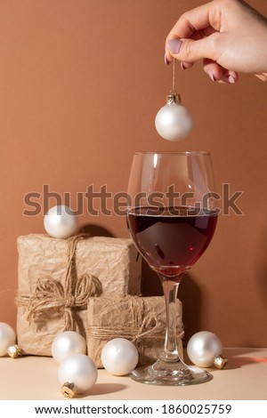 Hand with white christmas ball above the wineglass with red wine with paper gift boxes and white christmas balls background. Beige surface. Holiday concept. Festive evening. Holiday mood.