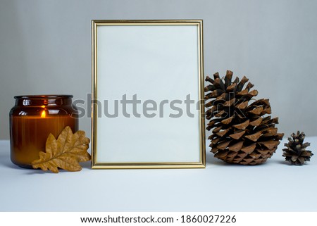Empty gold picture frame on white background. Mock up frame. A large pine cone, a candle, autumn leaves. Autumn empty frame. Autumn background. Autumn composition. Cozy house. Copy space. Decoration.