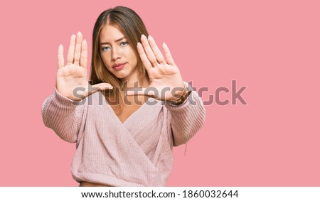 Beautiful blonde woman wearing casual winter pink sweater doing frame using hands palms and fingers, camera perspective 