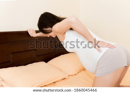 beautiful pregnant woman touching her tummy and keeping one hand on her back at home on bed.