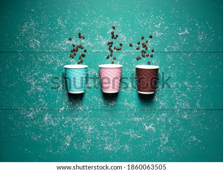 Multi-colored paper cups for coffee lie on a wooden turquoise background, strewn with snow.